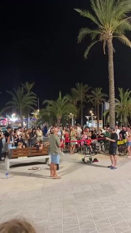 At night, a crowd gathers on a paved area in front of palm trees. Two men stand in the center of the paved area, facing each other. The man on the left is wearing a light-colored short-sleeved shirt and blue shorts. The man on the right is wearing a dark t-shirt and dark shorts. Between them, a red ribbon is stretched horizontally, and the man on the right is holding one end of it. In front of the two men, a small, wheeled platform with a red object on it is visible. A crowd of people of various ages stand around them, watching. Some people are sitting on a bench to the left. The atmosphere appears to be festive.