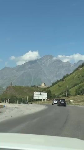 The video captures a scenic view from inside a vehicle, driving on a winding asphalt road through a mountainous landscape. The road curves to the right, with two dark-colored cars ahead, one directly in front and another slightly to the left. The environment is characterized by lush green, sloping hillsides on the right, dotted with trees and a few scattered buildings. On the left, the terrain also slopes, revealing more distant buildings and trees. A prominent white billboard stands on the right side of the road. Majestic, rocky mountains dominate the background, with some peaks partially obscured by clouds. The sky is a clear, bright blue with scattered white clouds. The overall impression is of a journey through a beautiful, rural mountain region.