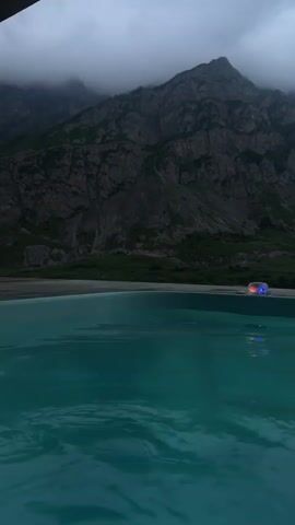 The video pans across a swimming pool with turquoise water in the foreground. In the middle ground, there is a dark tiled edge of the pool. In the background, a mountain range is visible under a cloudy sky. A small object with red and blue lights is floating on the surface of the water near the edge of the pool. The lighting suggests it is dusk or a heavily overcast day.