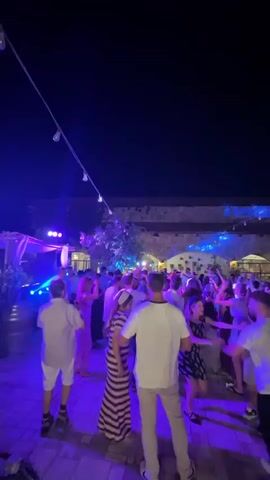 This is a vertical, low-angle, first-person perspective video taken at night. The scene is a crowded outdoor party or festival. The lighting is predominantly purple and blue, with some white lights visible. People are dancing and interacting. The ground appears to be paved with cobblestones or similar material. In the background, there are buildings with arched openings. Strings of lights are suspended overhead, adding to the festive atmosphere. The camera is moving, suggesting the videographer is walking through the crowd.