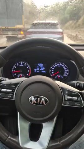 The video shows the interior of a car, looking through the windshield which is covered in raindrops. The steering wheel, dashboard, and a digital display are visible. In front of the car, there are other vehicles on a dirt road, including a truck and a car, all moving slowly. The weather appears to be rainy or wet, with the sky overcast. The scene suggests a slow-moving traffic situation in inclement weather on an unpaved road.