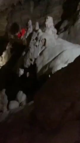 The video shows the inside of a cave, with various rock formations visible. The cave is dimly lit, with some areas appearing darker than others. Some of the formations have a smooth, white surface, while others are rough and gray. In some parts of the video, stalactites are visible hanging from the ceiling. A person wearing a bright red outfit is seen climbing on some of the higher rock formations. A metal railing is visible in the foreground.