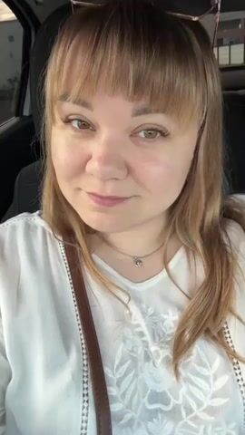A woman with blonde hair and bangs is in the driver's seat of a car. She is wearing a white top with embroidery and a brown purse strap is visible. She is looking at the camera and touching her hair. The background is out of focus.
