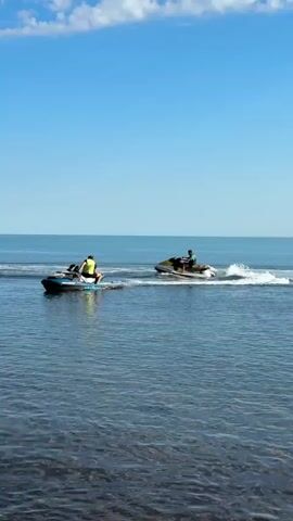 The video captures a bright daytime scene of two individuals riding jet skis on a calm, clear blue body of water. The setting is coastal, with a rocky and pebbled shore visible in the foreground, dotted with larger rocks and some dark marine vegetation. In the background, a distant coastline featuring green cliffs and trees stretches along the horizon.

One person, wearing a yellow or lime green life vest, is seated on a dark-colored jet ski positioned closer to the shore. The other person, also wearing a light-colored top or life vest, is riding a yellow and black jet ski further out in the water, creating a white wake behind it. Both jet skis are moving away from the camera and towards the open sea. The sky above is a clear, bright blue with a few scattered white clouds, indicating sunny weather. The overall atmosphere suggests leisure and outdoor recreation in Divnomorskoe, Russia.