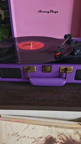 A turntable is playing a vinyl record. The turntable is purple with gold-colored latches and a handle. The lid is open and is light pink with the words "Sunny Days" printed on it. The record playing is black with a red center label. The turntable is placed on top of a wooden table. Underneath the table, at the bottom of the shot, are several vinyl record covers. The room also has a green potted plant in the background. The record is spinning and the needle is playing the music. The music has a strong, repetitive beat.