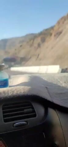 The video shows a view from inside a vehicle, looking out the windshield. In the foreground, on the dashboard, is a small glass vial with blue liquid and a silver cap. The vehicle appears to be on a highway that winds through a mountainous region. The road has multiple lanes and is bordered by a concrete barrier. Several trucks and other vehicles are visible on the road ahead. The mountains on either side of the road are dry and rocky, with sparse vegetation. The sky is clear and blue. There are billboards visible on the side of the road.