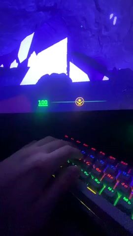 A first-person perspective video shows a person's hand playing a video game on a computer. The keyboard is illuminated with colorful lights. The screen shows a game, possibly Counter-Strike, with the player holding a knife and navigating a blue and white environment. The player's health is displayed as "100."