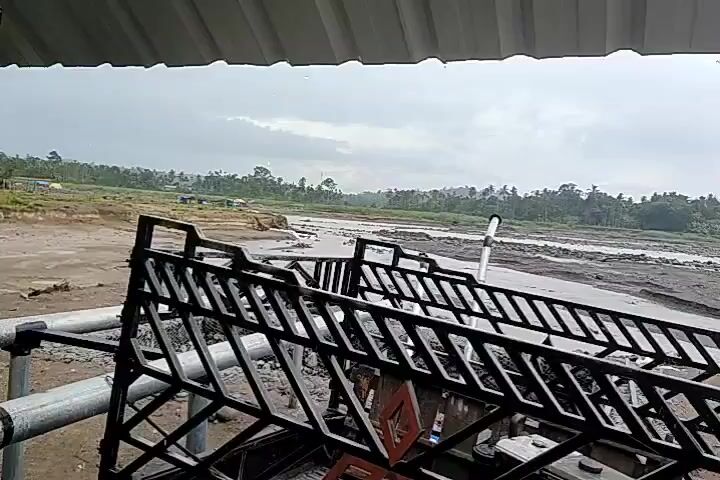 The video shows a muddy riverbed with a cloudy sky overhead. In the foreground, a black metal structure, possibly part of a vehicle or scaffolding, partially obscures the view. The river is wide and appears to be shallow in places, with visible muddy banks. In the middle ground, there are several yellow trucks and construction equipment on the riverbed, suggesting ongoing work or recovery efforts. The background consists of a treeline and distant hills. The overall impression is of a landscape affected by recent heavy rainfall or flooding, with a focus on the altered terrain and the presence of machinery.