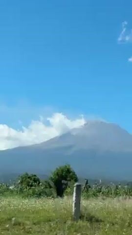 The video depicts a majestic mountain with its peak partially obscured by clouds. The sky is a vibrant blue, adding to the scenic beauty. In the foreground, lush greenery and trees provide a contrast to the mountain's grey hue. The camera appears to be moving, possibly from a vehicle on a road, capturing the landscape from a dynamic perspective.
