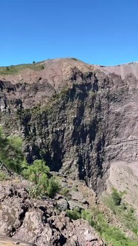 The video shows a vast, deep volcanic crater under a clear blue sky. The crater walls are rugged and layered, with visible signs of rock erosion and debris. Green vegetation, including bushes and grass, grows on the slopes and at the base of the crater. In the foreground, there are rocks and a wooden railing, suggesting a viewing platform. The camera pans slowly across the landscape, offering a panoramic view of the crater's immense scale and geological features.