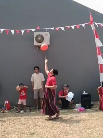 In an outdoor setting, a group of people are gathered in front of a gray wall decorated with red and white flags. A man in a red shirt and a sarong is playing with a red balloon, tossing it up in the air. He is barefoot. To his left, a young boy in a red shirt is holding a balloon. Behind them stands another man in a light-colored t-shirt and shorts. To the right of the man with the balloon, someone is partially visible behind a speaker. The ground is covered with gravel and dry grass. An air conditioning unit is mounted on the wall above the man with the balloon. The overall atmosphere suggests a casual event or celebration.