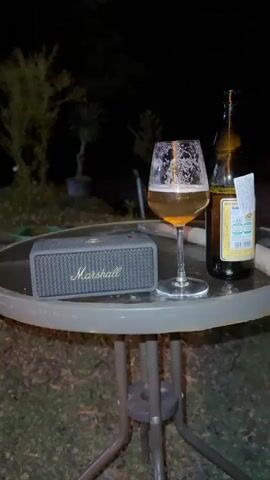 The video shows a dimly lit outdoor scene at night. A small, round metal table holds a Marshall Bluetooth speaker, a glass of light-colored beer with foam, and an open bottle of beer with a label. The table is set on a lawn or grassy area. A person is sitting in a chair, visible only from the knees down, wearing grey pants and white sneakers. The camera pans from the table to the person's legs and feet. The background is mostly dark with some blurry foliage visible.