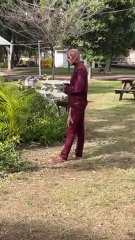 A man stands barefoot in a grassy, park-like setting. He is wearing a burgundy tracksuit with white stripes down the side, and has a bald head and beard. He appears to be putting on a pair of black gloves. Behind him are various bushes, plants, and a small building. There is also a picnic table visible in the background.