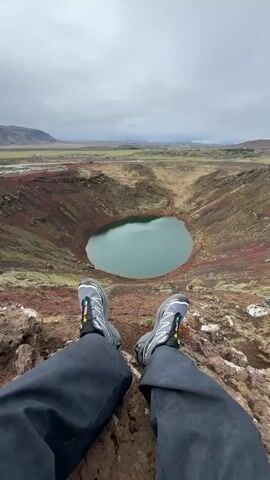 The video shows a person sitting on the edge of a volcanic crater, presumably in Iceland. The person is wearing dark pants and grey Salomon trail running shoes. The crater is a deep, reddish-brown color and contains a pool of turquoise water. The surrounding landscape is flat and grassy, with some buildings visible in the distance. The sky is overcast and grey.