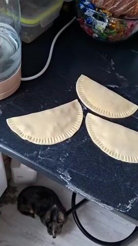 The video displays a dark, speckled countertop with several uncooked, crescent-shaped pastries arranged in rows. Each pastry has a crimped edge. In the background, on the countertop, are various items including bottles of what appear to be beverages, a carton with images of cherries, a bottle of cooking oil, and a plate of food. To the left, a small, dark-colored dog is visible on the floor, looking upwards. The lighting suggests an indoor environment, possibly a kitchen. The camera pans slightly to the right, showcasing more of the pastries and the items on the countertop.