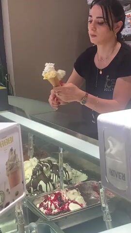 A woman wearing a black t-shirt with a logo is serving ice cream. She is holding an ice cream cone in her left hand, which has scoops of ice cream on it. She is using a scoop with her right hand to add more ice cream to the cone. In front of her is a display case filled with various flavors of ice cream. To her left, there is a sign with the word "BABBI" on it and an image of an ice cream cone. To her right, there is a white container with a red lid, and a dispenser for napkins. The background appears to be an ice cream shop, with shelves of snacks visible behind the woman. The lighting is warm and bright.