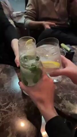 Here's a concise description of the video:

The video shows a group of people toasting with drinks in a bar setting. The drinks appear to be cocktails with ice, lemon slices, and mint. A woman then raises her drink and takes a selfie, smiling and laughing. The environment looks like a dimly lit bar with marble-patterned tables. A window with a multicolored reflective coating is visible in the background.