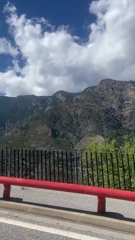 The video shows a view from a moving vehicle looking out at a mountainous landscape. A red railing and a black fence are visible in the foreground. The sky is blue with scattered white clouds. The mountains are covered in green trees and rocky terrain. A large tree is seen on the right side of the frame. The sun is shining brightly, casting shadows.