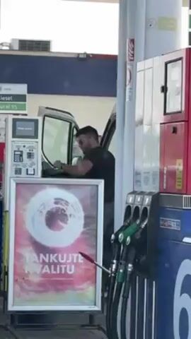 A medium shot depicts a gas station environment. A man with short dark hair, wearing sunglasses and a black t-shirt, is standing inside the open driver's side door of a white van. He's leaning on the steering wheel, looking down. To the left of the van is a gas pump kiosk with a screen and keypad. In front of the kiosk is an advertisement poster with the words "TANKUJTE KVALITU" (refuel quality) and an abstract design. To the right of the van are several gas pumps with green nozzles, next to a red metal utility box. The scene is brightly lit.