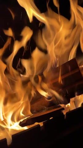 The video displays a close-up shot of a vibrant fire. Bright orange and yellow flames dance and flicker upwards from burning pieces of wood, which rest on a dark, metallic grate or frame. Sparks frequently fly upwards from the fire, creating a dynamic visual effect against the dark background. The wood appears to be in various stages of combustion, with some pieces still intact and others glowing red. The scene suggests a contained fire, possibly in a grill or fire pit, providing warmth and light in a dark setting. The audio features the crackling sounds of the burning wood, enhancing the immersive experience of the fire.