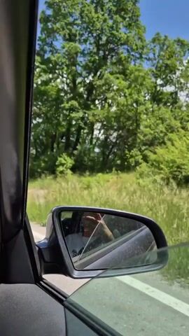 The video is filmed from the interior of a moving vehicle, specifically from the passenger side. It begins with a view of the side mirror, reflecting a hand holding a phone, then pans to the front windshield, revealing a road stretching into the distance through a rural landscape. The environment features green fields, scattered trees, distant hills, and a sky with white clouds. A car air freshener is visible on the dashboard.