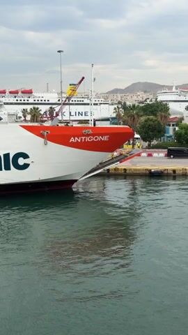 Here is a description of the video.

The video shows a large ferry boat called "ANTIGONE" docked at a harbor. The boat is white with a red bow and the name "saronic" in large blue letters on its side. The ramp at the front of the boat is extended and resting on the dock. Other large ships are visible in the background, along with palm trees and buildings on the shoreline. People can be seen walking on the dock near the boat. The water is calm, reflecting the boat and the sky, which is cloudy.