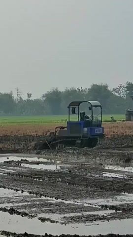 A compact blue tractor with a black cab and a canopy operates in a muddy field. The tractor is equipped with tracks and is working the soil, creating rows and disturbing the water. The field appears to be a paddy field, with evidence of recent cultivation. The background features a line of trees and a hazy sky. The overall scene depicts agricultural work in a rural setting.