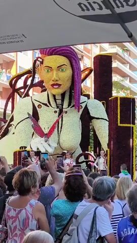The video shows a large, floral sculpture of a woman's head and torso, adorned with purple dreadlocks and wearing a white, futuristic outfit. A red and white butterfly is perched on her hand. She is on a float being pulled through a crowd of people, many of whom are holding up phones to take pictures and videos. The crowd is diverse in age and gender, dressed in casual summer clothing. Behind the float, buildings with balconies and windows are visible, suggesting an urban outdoor setting during daylight hours. Some individuals on the float appear to be performers or participants, dressed in white and interacting with the crowd. The overall atmosphere suggests a festive event or parade.