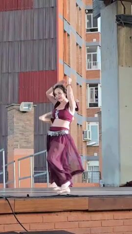A group of female dancers in maroon, sheer, flowing pants and matching crop tops with off-the-shoulder sleeves perform a coordinated dance on a wooden stage. The dancers have dark hair and are barefoot. Behind them is a building with orange and grey panels and several windows. The lighting appears to be natural daylight. One dancer in the foreground is facing right, with her arms extended above her head. Another dancer is visible behind her, also facing right. The dance appears to be a form of belly dancing.