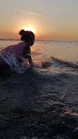 The video shows a young person running into the ocean at sunset. The sun is a bright orb on the horizon, casting a warm glow on the water. The person is wearing a purple shirt and dark shorts. They are kicking up a spray of water as they run, and their hair is pulled back in a bun. The waves are gentle, and the overall scene is peaceful and serene.