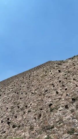 The video shows different areas of the archeological site Teotihuacan in Mexico. The first shot shows the side of a large stone pyramid under a clear blue sky. The camera then pans left, showing the pyramid's steps and a dirt path next to it. In the distance, a smaller pyramid is visible on a small hill surrounded by trees. A group of people is walking across a bridge or path, and a sign saying "Se Prohibe" (Forbidden) in Spanish is also visible. The video ends with a shot of white puffy clouds against a blue sky, with the tops of some trees visible in the lower part of the frame.