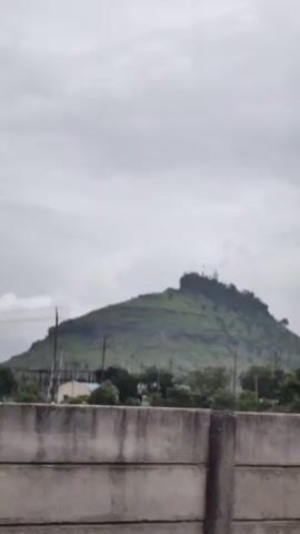 The video shows a cloudy day with a green, mountainous landscape. A concrete wall runs horizontally across the lower part of the frame. Trees and a few structures are visible at the base of the mountain. The peak of the mountain is flat and appears to have some buildings or structures on it. The video is shot from a low angle, looking up towards the mountain. The camera pans slightly to the left and then back to the right.