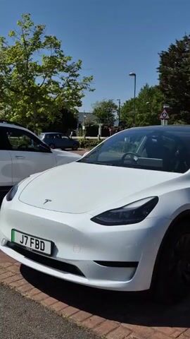 The video shows a white Tesla Model Y parked in a parking lot. The car has a black panoramic roof and black wheels. There is a green-accented license plate on the front of the car with the registration number "J7 FBD". Other cars are parked nearby, and trees and street lights are visible in the background. The sky is blue, and it appears to be a sunny day. The camera pans around the front of the car to provide a better view of the vehicle.