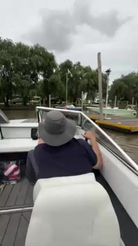 From a camera inside a boat, a man wearing a hat and dark shirt sits in the driver's seat and steers the boat away from a dock on an overcast day. The boat is white with grey flooring. In the foreground, a white seat faces forward. Through the windshield, a view of the dock and surrounding green trees is visible. There's a bag with a red and white cloth in the background.