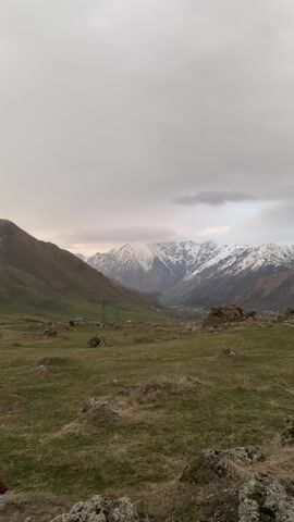 The video shows a landscape view of mountains under a cloudy sky. In the foreground, there's a field of green grass dotted with rocks. To the left and right, hills rise up, leading to a valley where power lines and what appear to be small buildings are visible. In the distance, the backdrop is dominated by snow-covered mountains partially obscured by clouds. The overall lighting is soft, suggesting it might be dawn or dusk.