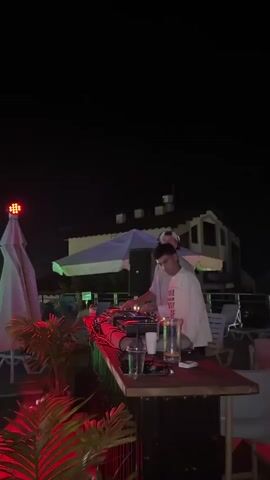 A nighttime scene unfolds on what appears to be a rooftop or elevated outdoor venue. In the foreground, a DJ booth is set up with two individuals attending to the equipment. Red and green lights illuminate the area, casting a vibrant glow on plants and furniture. Several people are visible in the midground, some standing and conversing, while others are near a swimming pool that glows with green light. A large white umbrella is prominent in the background, and a building with multiple windows is visible behind it. The overall atmosphere suggests a social gathering or party.