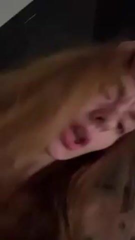 A close-up, low-angle shot features a person with long, light brown hair that partially obscures their face. The person’s lips are slightly parted, and they appear to be sniffling or congested. The background is dark and blurry, with a glimpse of what seems to be a dark surface. The camera angle is unsteady, suggesting a handheld recording.