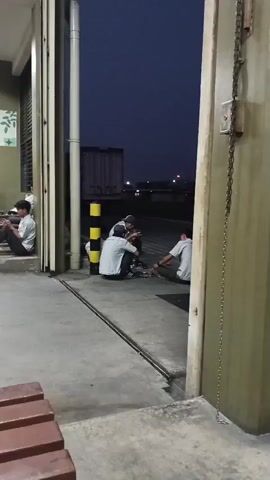 The video shows an outdoor scene at dusk or night. In the foreground, there is a concrete ground and a wooden bench. To the left, a person in a white shirt and dark pants is sitting. In the middle ground, a group of three people, all wearing white shirts and dark pants, are sitting on the ground. One person is holding something in their hands, and there are some objects on the ground in front of them. To the right, there is a large industrial door or building facade with a chain hanging down. In the background, there is a large white shipping container and some distant lights, suggesting an industrial or port area. The sky is dark blue.