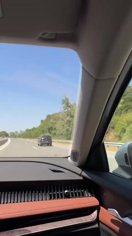 The video is taken from inside a car, looking out the windshield and side window. The car is driving on a highway with a clear blue sky and some clouds. Trees and vegetation line the side of the road, with a hill in the background. Another car is visible ahead on the highway. The interior of the car features a brown dashboard and trim. The camera appears to be stationary, capturing the view from the passenger seat. The audio is music.