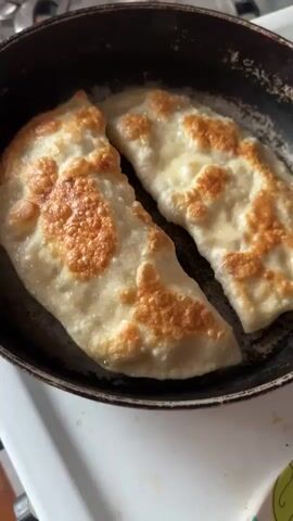 The video shows two chebureki frying in a pan. The chebureki are golden brown and puffy, with some areas of darker browning. The pan is black and appears to be well-seasoned. The chebureki are arranged side by side in the pan, and there is some oil visible around them. The background is out of focus, but it appears to be a kitchen setting.