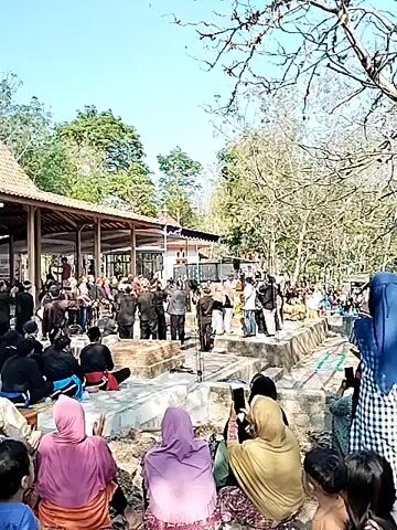 A crowd of people are gathered outdoors. In the foreground, several people are seated, facing away from the camera, with some wearing headscarves. Behind them, a group of people stand in an area that appears to be a shrine or burial site. Some individuals in this group are wearing traditional clothing. A microphone is visible on a stand in front of the standing group. In the background, a large, bare tree dominates the frame, with more people visible around its base. The sky is clear and blue. The overall atmosphere suggests a gathering or ceremony.