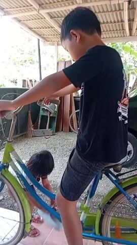 A young boy in a black t-shirt and shorts is sitting on a bicycle, with his left foot on the pedal and his right foot resting on the ground. Another young child is crouched down near the front wheel of the bicycle. The scene appears to be outdoors, possibly in a yard or alleyway, with a corrugated metal roof overhead and some stored items in the background. The boy is holding the handlebars of the bicycle.