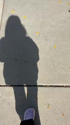 The video captures a first-person perspective of a person walking on a concrete sidewalk in Seal Beach, USA, during daylight. The person's shadow is cast prominently in front of them, indicating a sunny day. The shadow of their left hand is visibly making a "peace sign" gesture. The person's right foot, clad in a light purple Croc shoe and black pants, is visible in the lower right portion of the frame, moving forward. Small, bright yellow and orange leaves are scattered across the grey concrete ground. The scene suggests a casual walk on a clear, sunny day.