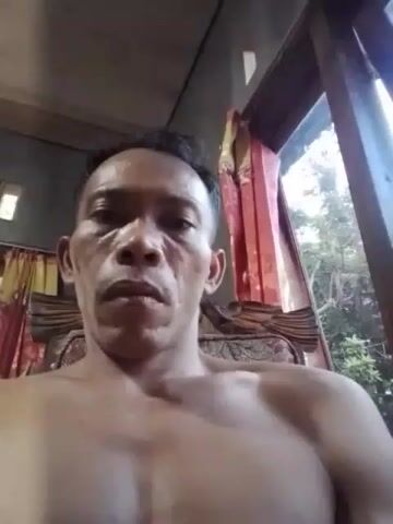 The video shows a close-up, selfie-style shot of a shirtless man. He appears to be indoors, with curtains and a window visible in the background, suggesting a residential setting. The man's face is the primary focus, and he is looking directly at the camera. The lighting suggests it is daytime, possibly with natural light coming from the window. There are no other people or significant objects in the frame besides the man and the immediate surroundings.