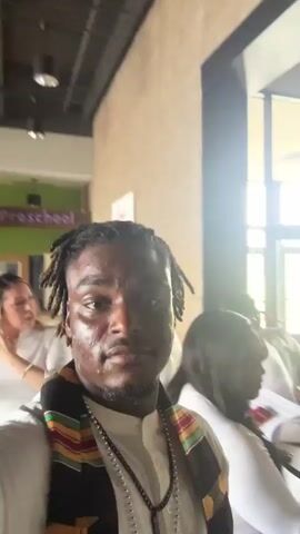 A man with dreadlocks wearing a white shirt and a colorful patterned scarf or stole records a selfie video, smiling and appearing cheerful. He is positioned in the foreground, facing the camera. In the background, several women are visible; most are wearing white attire, suggesting a possible uniform or coordinated dress code. They are walking or standing in what appears to be a hallway or lobby of a building.

The setting features light brown brick walls and large windows letting in natural light, indicating it is daytime. Overhead, modern hanging light fixtures are visible. In the background, a sign with "Preschool" written on it is clearly visible, suggesting the location is a school or a facility with a children's area. Another white sign on the brick wall has partially readable text, which appears to say "love God. love people." A potted plant is also seen in the background.