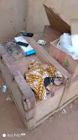 The camera pans around a messy outdoor area. The camera starts focused on an old wooden cot with various items strewn on it: a phone, a phone box, and a colorful piece of fabric. The camera then pans right, showing wooden boxes and pieces of wood. It then pans right again to a view of a metal gate that leads to an open, sandy area with some distant structures. Finally, the camera pans back to the wooden cot. The scene appears to be somewhat neglected and utilitarian. The recording device appears to be an itel P40 phone.
