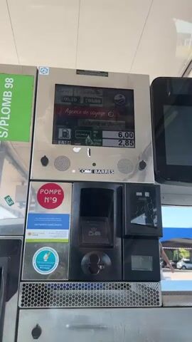 The video shows a close-up of a gas pump at a station. The screen displays information about gas prices and operating hours. The pump has a card reader and a slot for bills. There is a person standing in the background, but they are out of focus and their actions are not clear. The environment appears to be an outdoor gas station with other pumps visible in the background.