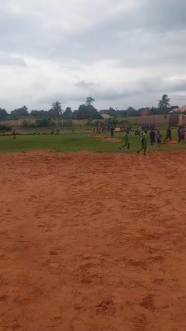 The video captures a group of children playing on a grassy field with a dirt area in the foreground. In the distance, buildings are visible, along with trees and a fence. The sky is cloudy. Some children are kicking a ball, while others are running and interacting. One child in the foreground walks away from the camera.