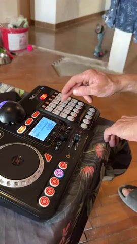 A pair of hands operate a black electronic musical instrument with buttons and a screen, resting on a table covered with a patterned cloth. The background shows a floor with a pattern and a partial view of a wall. One hand is seen pressing buttons on the instrument.