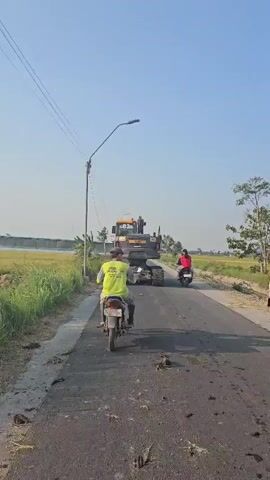 The video shows a rear view of a motorcycle with a rider wearing a neon yellow shirt. The motorcycle is on an asphalt road. Ahead of the motorcycle is a large yellow construction vehicle, likely a paver, with a trailer. The construction vehicle appears to be moving forward on the road. To the right of the road are dry grass and trees, and to the left are green grass and a body of water. The sky is clear and blue. Another motorcycle with a rider is visible in the distance, ahead of the construction vehicle.
